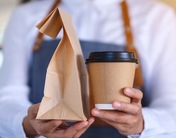 Primer plano de una camarera sosteniendo y sirviendo un café en envase para llevar y comida en una bolsa de papel.